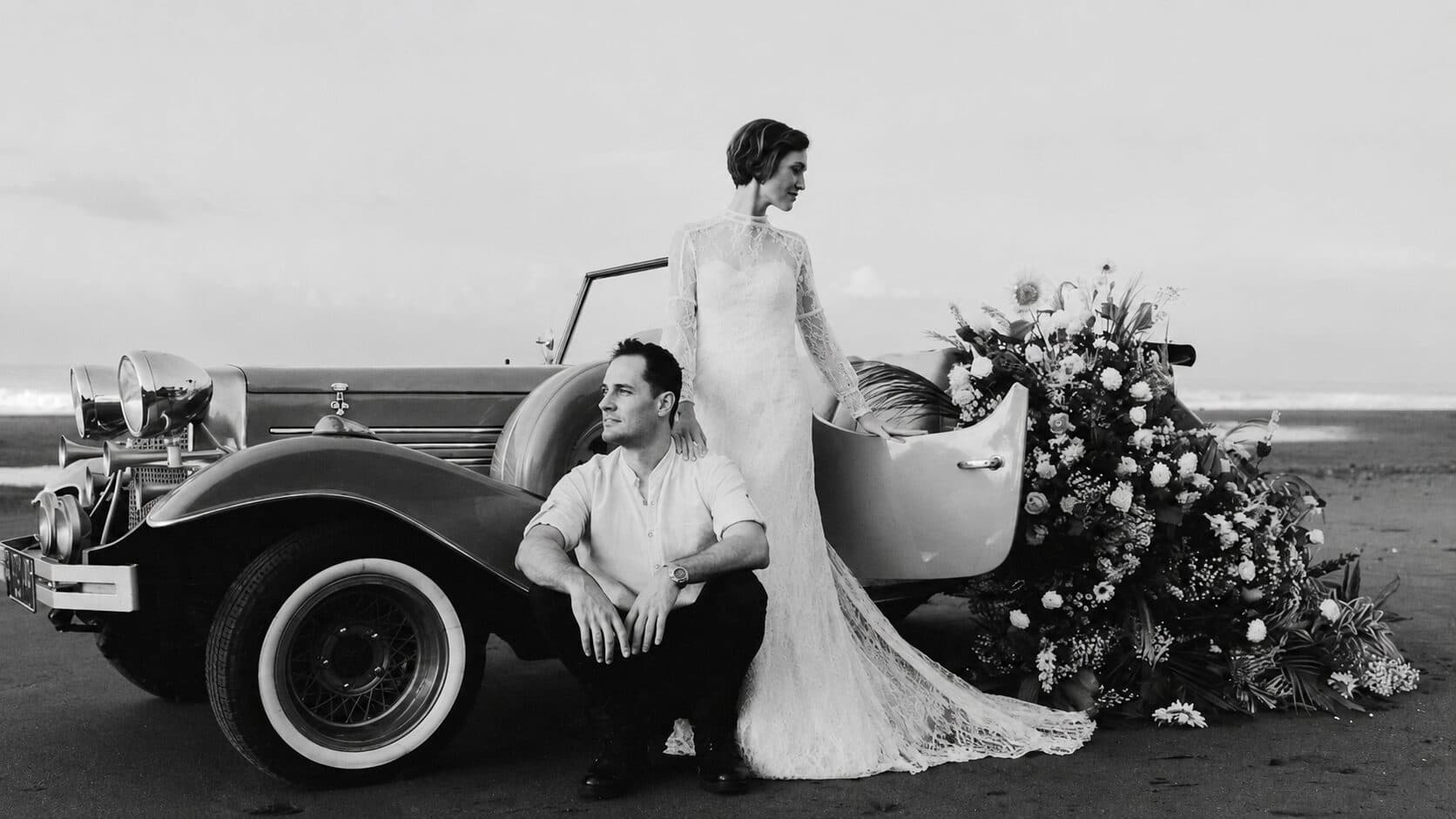 A couple beside a vintage car on a Bali beach, with a cascading floral arrangement