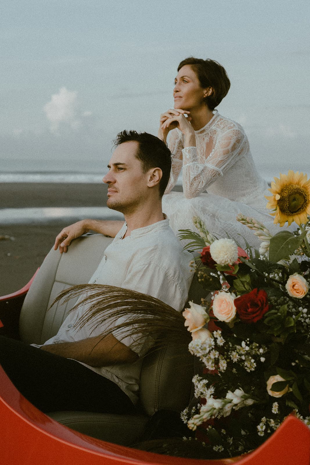 A couple seated in a vintage red car with wedding florals by the beach