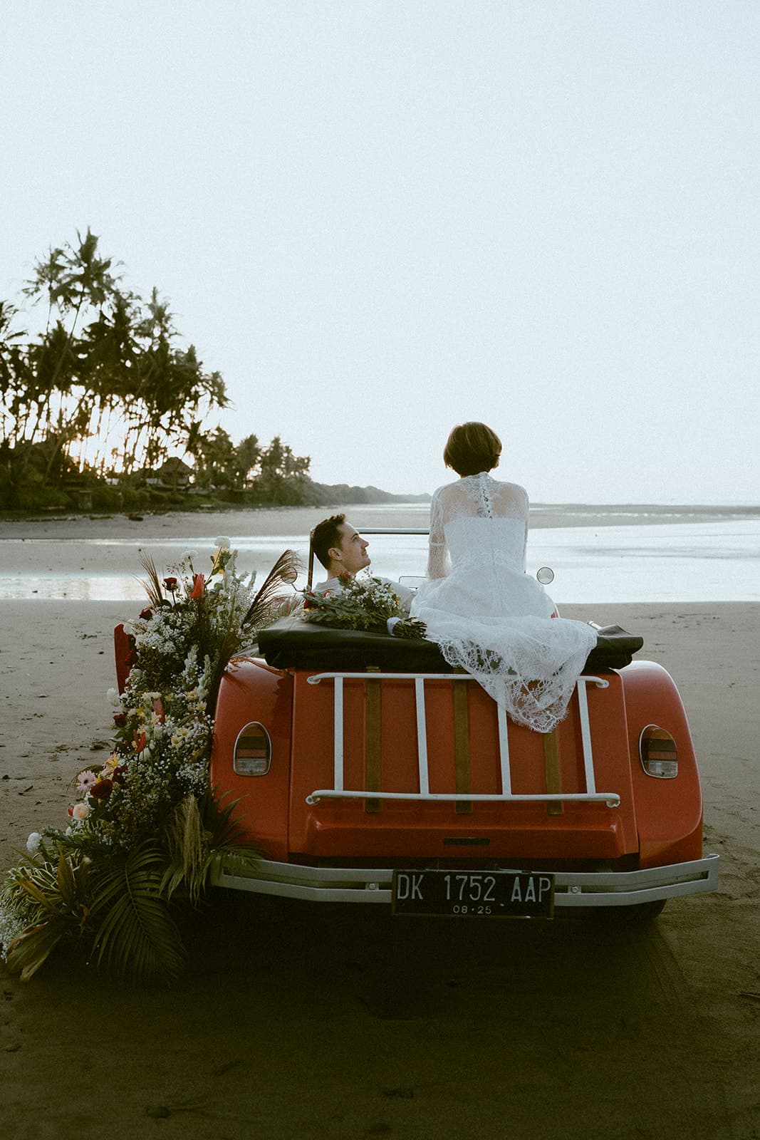 A couple seated on a vintage red car overlooking a beach at sunset