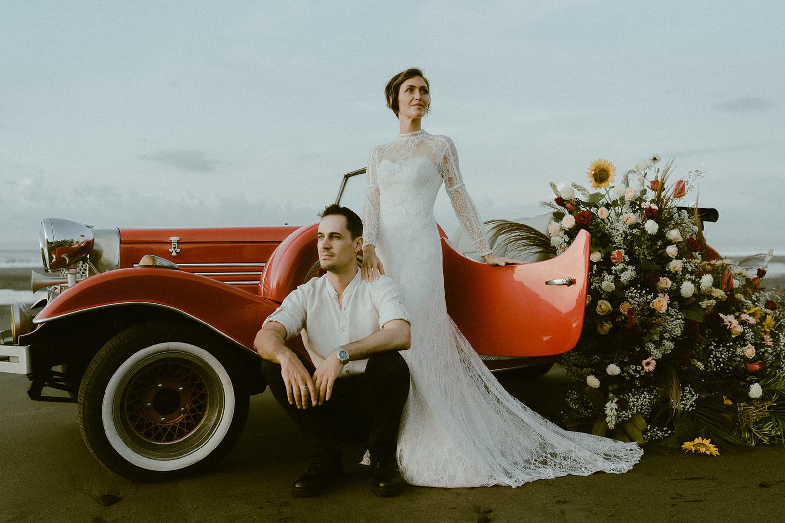 A newlywed couple by a vintage red car on the beach