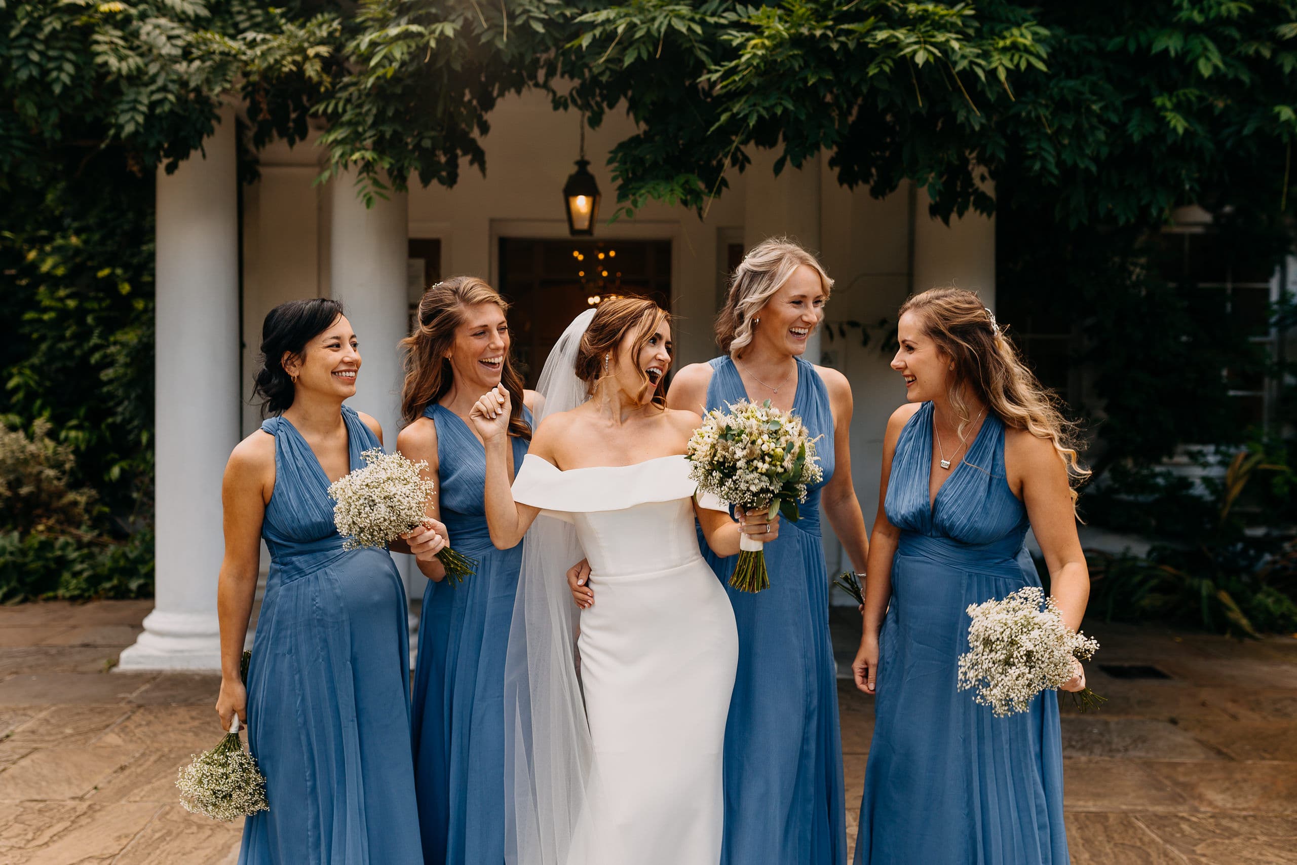 A bride laughing with her bridesmaids in blue dresses outside a wedding venue